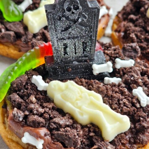 These Mini Graveyard Cookie Cakes turn refrigerated chocolate-chip cookie dough into little Halloween graveyards! Each cookie cake gets a layer of chocolate frosting, crushed Oreo “dirt,” and chocolate tombstones for the graveyard and white chocolate skeletons, plus a gummy worm!