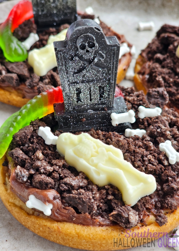 These Mini Graveyard Cookie Cakes turn refrigerated chocolate-chip cookie dough into little Halloween graveyards! Each cookie cake gets a layer of chocolate frosting, crushed Oreo “dirt,” and chocolate tombstones for the graveyard and white chocolate skeletons, plus a gummy worm!